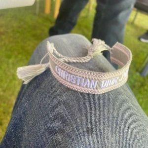 Beige armband från Christian Dior - Säljer ett snyggt beige armband från Christian Dior med broderad logga i vitt. Armbandet har justerbar knytning med tofsar i ändarna och är gjort i ett mjukt tygmaterial. Perfekt accessoar för att lyfta din outfit.