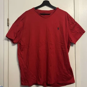 Röd t-shirt från Polo Ralph Lauren - Röd t-shirt från Polo Ralph Lauren med klassisk v-ringning och broderad marinblå logga på bröstet. Tillverkad i 100% bomull för en skön och luftig känsla. Perfekt basic-plagg med normal passform och korta ärmar.