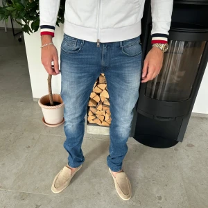 Replay Anbass jeans  - Snygga Replay Anbass jeans i klassisk blå tvätt med diskret slitning. Modellen har normal passform, raka ben och fem fickor. Tydliga Replay-detaljer på bakfickorna och läderpatch i midjan. Perfekta för en avslappnad och trendig look.