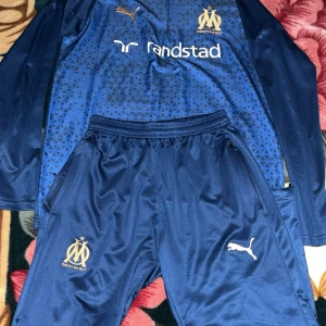 Marinblå Marseille fotbollströja Puma - Snygg marinblå långärmad fotbollströja från Puma för Olympique de Marseille. Tröjan har geometriskt mönster, klubbmärke och sponsortryck på bröstet. Tillverkad i glansigt funktionsmaterial, perfekt för träning eller match.
