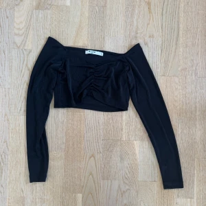 Svart offshoulder topp från NA-KD - Trendig svart croppad topp från NA-KD med offshoulder-design och långa ärmar. Toppen har snygg rynkdetalj framtill som ger en cool look. Perfekt att styla med höga jeans eller kjol för en modern vibe.