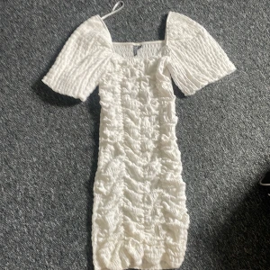 Vit kort klänning med puffärm - Supersöt vit kort klänning med puffiga kortärmar och smockad struktur. Klänningen har fyrkantig ringning och sitter tight över kroppen. Perfekt för sommarens alla tillfällen och ger en fräsch och trendig look.