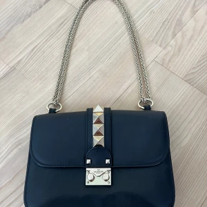 Svart axelväska Valentino med gulddetaljer - Snygg svart axelväska från Valentino Garavani i skinn med kedjerem i guld och ikoniska pyramidformade guldfärgade nitar på locket. Väskan har ett elegant lås framtill och stilren design som lyfter varje outfit.