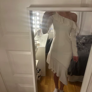 Vit asymmetrisk klänning med volang - Supersnygg vit klänning med asymmetrisk design och volangdetalj längs axeln och nederkanten. Ena axeln är bar och den andra har lång ärm, vilket ger en unik och trendig look. Perfekt för dig som vill sticka ut med en elegant och modern stil.