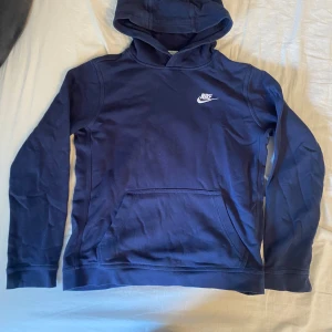 Mörkblå Nike hoodie med huva XL - Snygg mörkblå hoodie från Nike med klassisk logga på bröstet. Tröjan har huva, känguruficka och långa ärmar. Perfekt för chill dagar eller när du vill ha en sportig look. Tillverkad i mjuk bomullsmix för extra komfort.