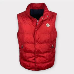 Äkta Moncler väst  - Snygg röd dunväst från Moncler med hög krage och klassisk logga på bröstet. Västen har två fickor med dragkedja och knäppning framtill. Insidan har ett coolt serietryck. Storleken är 6 vilket blir som en L-XL. Bilden på insidan har lossnat lite på översidan vilket gör att priset är sänkt. Äkta såklart. Kom gärna med bud.