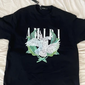 Svart Amiri t-shirt med fågeltryck - Svart t-shirt från Amiri med stort grafiskt tryck framtill som visar två vita fåglar och gröna blad. Klassisk rund halsringning och korta ärmar. T-shirten är i mjuk bomull och har en modern, avslappnad vibe.