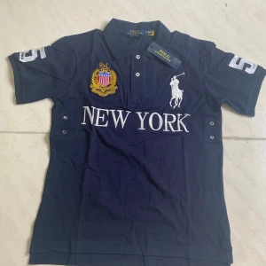  Polo Ralph Lauren Chief Keef - Snygg marinblå t-shirt från Polo Ralph Lauren med broderad logga, stort 'NEW YORK'-tryck och emblem framtill. Klassisk pikékrage, knappar och stora siffran 5 på ryggen. Skön bomullskvalitet och sportig vibe, perfekt för dig som gillar streetstyle.