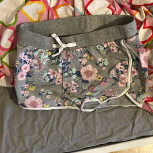 Grå blommiga shorts från Ellos - Superfina grå shorts från Ellos med blommigt mönster i rosa, blått och gult. De har vit snörning i midjan och vita kanter längs benen. Perfekta för sommardagar eller chill hemma. Mjukt bomullsmaterial och avslappnad passform.