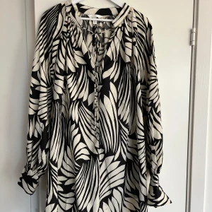 Svartvit mönstrad klänning H&M - Snygg svartvit klänning från H&M med grafiskt mönster, långa ärmar och knytning vid halsen. Luftig modell i mjukt material, perfekt för dig som gillar statement-plagg med coola detaljer.