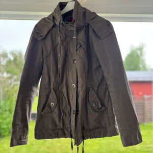 Olivgrön parkajacka från Vero Moda - Snygg olivgrön parkajacka från Vero Moda med huva, dragsko i midjan och stora fickor framtill. Jackan har långa ärmar, knappar och dragkedja framtill samt coola detaljer vid axlarna. Perfekt för dig som gillar en avslappnad och trendig stil.