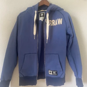 Blå hoodie från G-Star RAW med huva - Blå hoodie från G-Star RAW med vit broderad logga på bröstet, dragkedja framtill och stora fickor. Tröjan har vit snörning i huvan och ribbade muddar. Perfekt för dig som gillar streetwear och vill ha en chill look.