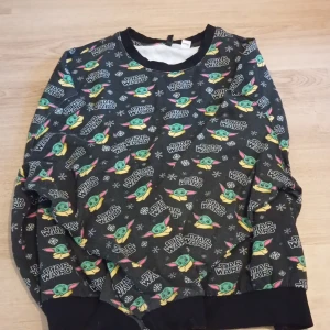 Star Wars tröja med Baby Yoda-motiv - Svart långärmad tröja med all-over print av Baby Yoda och Star Wars-logga. Tröjan har snöflingor i mönstret och ribbade muddar vid ärmslut och nederkant. Perfekt för dig som älskar Star Wars och vill ha en mysig och cool look.