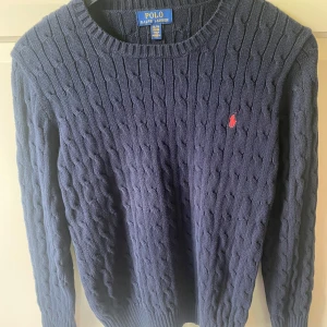 Marinblå kabelstickad tröja Polo Ralph Lauren - Kabelstickad marinblå tröja från Polo Ralph Lauren i storlek XL (18-20). Skulle säga att den passar S.  Klassisk rund halsringning och lång ärm, med röd broderad logga på bröstet. Tröjan är stickad i bomull och har ribbade muddar vid ärmslut och nederkant.