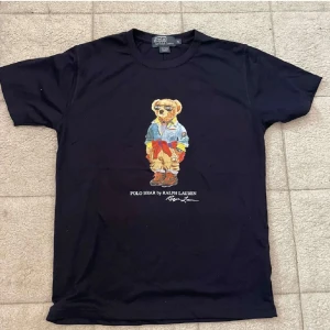 Mörkblå Polo Bear t-shirt från Ralph Lauren - Svart t-shirt från Polo Ralph Lauren med ikoniskt Polo Bear-tryck framtill. Klassisk passform med rund hals och korta ärmar. Tillverkad i 100% bomull för en skön känsla. Perfekt statement-plagg för dig som gillar streetstyle och snygga prints.