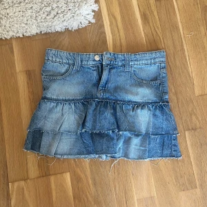 Blå jeanskjol med volanger - Snygg blå jeanskjol med två lager volanger och råa kanter för en cool, avslappnad look. Kjolen har klassiska fickor fram och bak samt hällor för bälte. Perfekt för dig som gillar denim och vill sticka ut lite extra. Helt ny!!! Storlek 158!