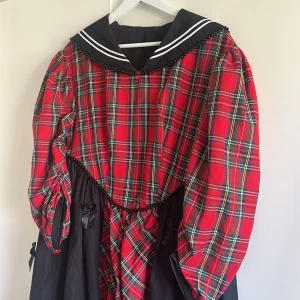 Rutig långklänning med sjömanskrage - Unik långklänning med röd och grön rutig överdel, svarta partier och dekorativa rosetter. Klänningen har puffiga ärmar, svart sjömanskrage med vita ränder och pärldetaljer längs kragen. Perfekt för dig som vill sticka ut med en cool vintagevibe.