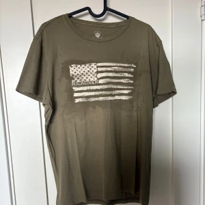 Olivgrön t-shirt från Ralph Lauren - Olivgrön t-shirt från Ralph Lauren med ett coolt, slitet tryck av den amerikanska flaggan på bröstet. Klassisk passform med rund hals och korta ärmar. Perfekt för en avslappnad streetstyle-look.