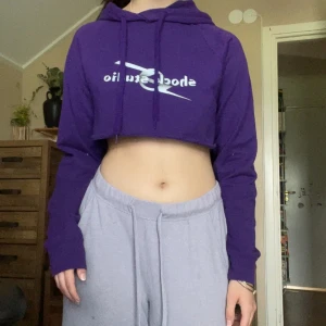 Lila croppad hoodie med tryck - Säljer en lila croppad hoodie från Fruit of the Loom med trycket 'Shock Studio' på bröstet. Tröjan har huva med snörning och är långärmad. Perfekt för dig som gillar streetstyle och vill ha en chill vibe.