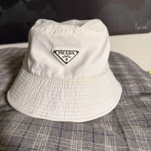 Vit bucket hat från Prada - Snygg vit bucket hat från Prada som inte kommer till användning för mig mera. Hatten har en stilren design med bred, nedåtlutande brätte och är tillverkad i ett slätt tyg som ger en clean look. Perfekt accessoar för att lyfta din outfit. Skriv gärna ifall du har mer frågor eller vill ha fler bilder!