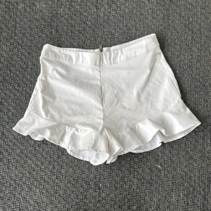 Vita volangshorts från Zara - Snygga vita shorts från Zara med volangkant nedtill och dragkedja bak. De är högmidjade och har en feminin, flowy look som passar perfekt till varma dagar. Materialet är mjukt och känns skönt mot huden.