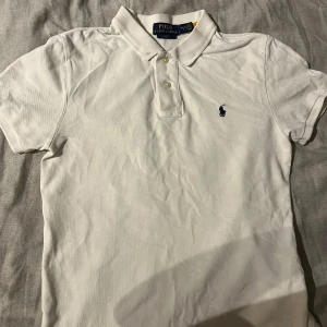 Vit pikétröja från Polo Ralph Lauren - Klassisk vit pikétröja från Polo Ralph Lauren med korta ärmar och broderad marinblå logga på bröstet. Tröjan har krage, två knappar framtill och är gjord i mjuk bomull. Perfekt för en clean och stilren look.