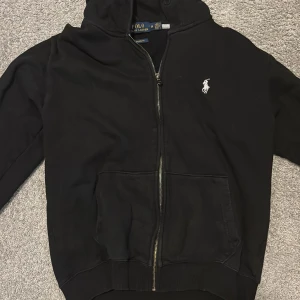 Svart hoodie från Polo Ralph Lauren - Svart hoodie från Polo Ralph Lauren med dragkedja och klassisk vit logga på bröstet. Tröjan har huva, känguruficka och ribbade muddar. Snygg och enkel design som funkar till det mesta. Custom fit-modell.