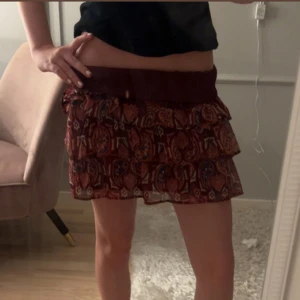 Vinröd volangkjol från Lindex - Supersöt kort volangkjol från Lindex i vinrött med färgglatt mönster i orange, blått och vitt. Kjolen har flera lager volanger och bred resår i midjan för skön passform. Perfekt för dig som gillar boho-vibe och vill sticka ut lite extra.