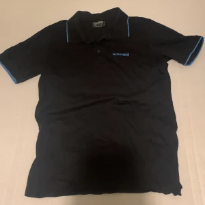 Svart pikétröja från McKenzie - Svart pikétröja från McKenzie med blå detaljer på krage och ärmslut. Klassisk pikékrage med knappar framtill och diskret logga på bröstet. Tillverkad i mjuk bomull som känns skön mot huden. Perfekt för en clean och sportig stil.