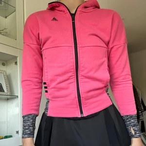 Rosa hoodiejacka från Adidas - Snygg rosa hoodiejacka från Adidas med dragkedja framtill och svart logga på bröstet. Jackan har huva, långa ärmar och svarta detaljer på sidorna. Perfekt för en sportig look eller till träning.