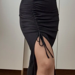 Svart långkjol med slits och snörning  - Svart långkjol i stretchigt material med snygg slits på sidan och trendig snörning som ger en rynkad effekt. Kjolen har en tight passform och är perfekt för dig som gillar stilrena plagg med en twist.