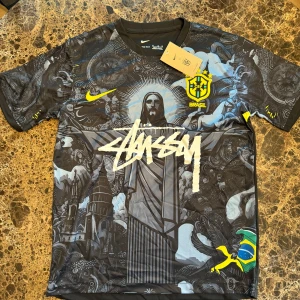 Nike Brasil x Stüssy svart fotbollströja M - Unik svart Brasil-fotbollströja från Nike och Stüssy med detaljerat grafiskt tryck, gult Brasil-märke och Stüssy-logga framtill. Tillverkad i Dri-FIT-material som andas. Korta ärmar med coola motiv och brasiliansk flagga på ärmarna.