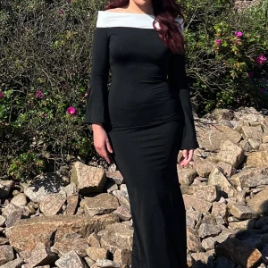 Svart långklänning med vit offshoulder - Elegant svart långklänning med vit offshoulder-detalj. Klänningen har långa ärmar med utsvängda ärmslut och en figurnära passform som framhäver siluetten. Perfekt för dig som vill sticka ut med en stilren och sofistikerad look.