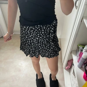 Svart blommig omlottkjol från Zara - Supersöt svart kort omlottshortskjol från Zara med små vita blommor och volangkant. Storlek S.