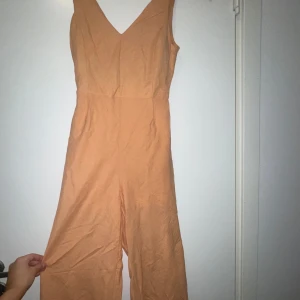 Jumpsuit  - Snygg persikofärgad jumpsuit med v-ringning och breda axelband. Byxdressen har en öppen rygg med knytband och vida ben. Tillverkad i ett mjukt bomullsliknande material, perfekt för varma dagar. Har liten skada på sidan som jag befogat bilf på:)