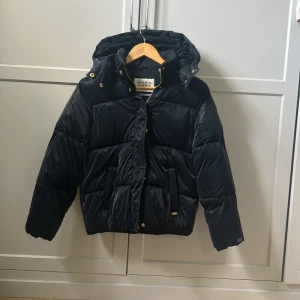 Mörkblå pufferjacka från Scotch & Soda - Svart pufferjacka från Scotch & Soda med huva och guldfärgade detaljer. Jackan har quiltad design, dragkedja och tryckknappar framtill. Perfekt för kalla dagar och har en cool, lite glansig finish som sticker ut. Den är oversized, storlek 152 men passar som XXS 