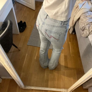 Ljusblå bootcut jeans med slitna knän - Säljer ett par ljusblå bootcut jeans med slitningar vid knäna och snygga röda detaljer på bakfickorna. Jeansen har låg midja och klassisk femficksdesign. Perfekta för dig som gillar en avslappnad och trendig look. Finns en fläck vid låret men syns knappast