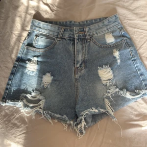 Blå slitna jeansshorts från SHEIN - Trendiga blå jeansshorts från SHEIN med hög midja och snygga slitningar framtill och baktill. Råa kanter nertill för en edgy vibe. Klassisk femficksmodell i 85% bomull och 15% polyester. Perfekta för sommardagar och festivaler.