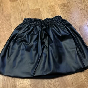 Svart kort kjol i skinnimitation H&M - Snygg svart kort kjol från H&M i glansig skinnimitation med bred resår i midjan. Kjolen har en lätt utställd form och är perfekt för dig som gillar en edgy look. Enkel att matcha med både t-shirt och stickad tröja.