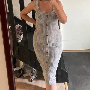 Grå ribbad gravid-klänning - Snygg grå ribbad gravid-klänning med knappar framtill. Klänningen är ärmlös, har en tight passform och går ner till knäna. 