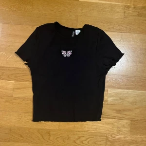 Svart croppad topp med fjäril - Svart croppad topp från Divided med en liten broderad fjäril i rosa och vitt på bröstet. T-shirten har korta ärmar och lätt vågig kant nertill för en extra cool vibe. Perfekt att matcha med jeans eller kjol.