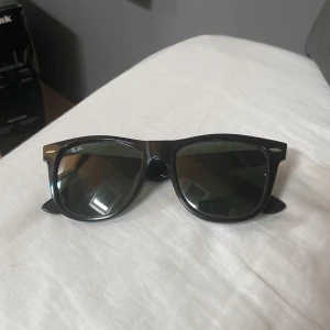 Svarta Ray-Ban Wayfarer solglasögon - Klassiska svarta Ray-Ban Wayfarer solglasögon med snygg, tidlös design. Bågarna är i blank svart plast och har gröntonade glas. Ray-Ban-loggan syns på både linsen och skalmen.  Du får med ett rayban fodral också