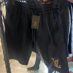 Svarta shorts från Louis Vuitton - FINNS ALLA STORLEKAR ! Svarta shorts från Louis Vuitton med guldfärgad LV-logga på ena benet. De har elastisk midja med snörning och två sidofickor. Perfekta för en avslappnad och trendig look.