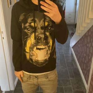 Svart hoodie från Givenchy med hundmotiv - Säljer en svart hoodie från Givenchy med ett stort tryck av en hund på framsidan. Hoodien är tillverkad i bomull och elastan för en bekväm passform. Perfekt för dig som vill ha en unik och stilren look.
