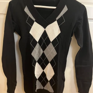 Svart tröja med diamantmönster - Snygg svart tröja med ett klassiskt diamantmönster i vitt och grått på framsidan. Tröjan har en v-ringad hals och långa ärmar, perfekt för en stilren look. Passar bra till både jeans och kjol.