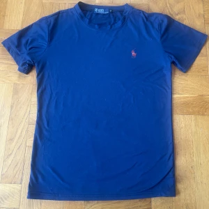 Mörkblå t-shirt från Ralph Lauren - Snygg mörkblå t-shirt från Ralph Lauren med en liten röd broderad logga på bröstet. Perfekt för en stilren look. Tillverkad i mjuk bomull och har korta ärmar. Mycket gott skick. Storlek M