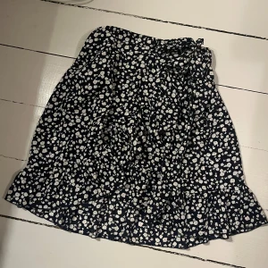 Blommig kjol med volang - Söt blommig kjol i svart och vita blommor med volangdetaljer. Kjolen har en omlott-design och en rosett i midjan. Storlek S. 🖤🤍