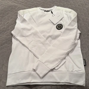 Vit sweatshirt från Balmain - Säljer en stilren vit sweatshirt från Balmain med ett svart emblem på bröstet och Balmain-logga längs ärmen. Tröjan har långa ärmar och rund halsringning. Perfekt för en avslappnad men ändå lyxig look.