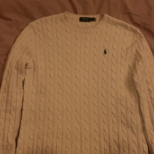 Vit kabel stickad tröja från Ralph Lauren - Säljer en klassisk vit stickad tröja från Ralph Lauren med kabelstickat mönster och den ikoniska loggan broderad på bröstet. Tröjan har långa ärmar och rund halsringning. Perfekt för en stilren look.