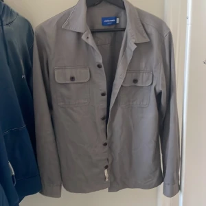 Grå jacka från Jack & Jones - Säljer en stilren grå jacka från Jack & Jones. Jackan har långa ärmar och knappar framtill samt två bröstfickor med knappar. Perfekt för en avslappnad look.